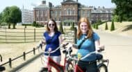 Two women pose with their bikes in front of Kensington Palace in London, England