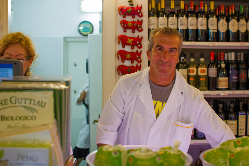 A smiling male market vendor standing in front of a shelf of wine in Italy